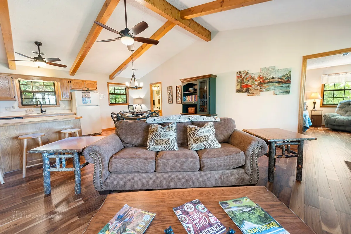 An inviting open-plan space combining a comfortable sofa with decorative pillows, a wooden dining table ready for shared meals or board games, and a glimpse into a fully equipped kitchen. Natural wood accents and vibrant wall art complete the cozy, welcoming atmosphere.