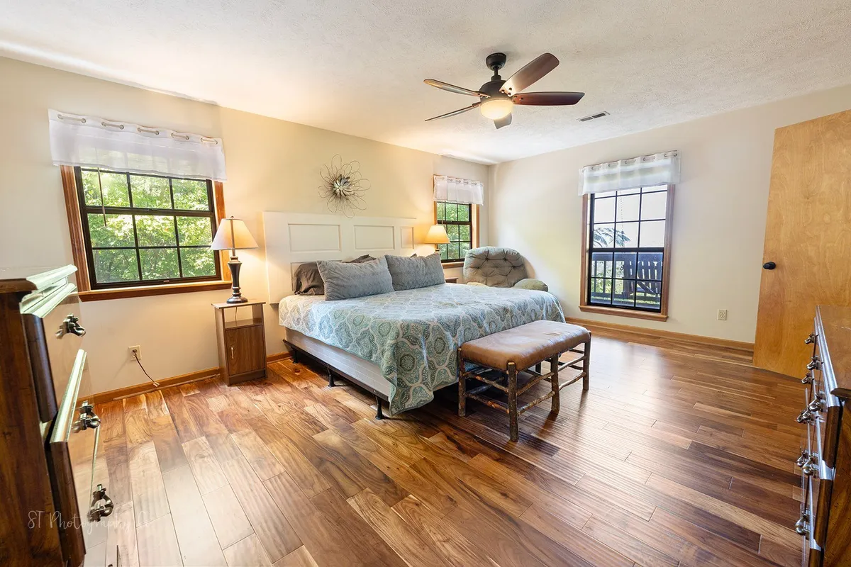 A bright and tranquil king bedroom featuring a large bed, windows letting in natural light, a dressing table, and a cozy bench at the foot of the bed. The room is decorated in warm, restful tones, inviting guests to unwind and enjoy peaceful mornings or evenings.