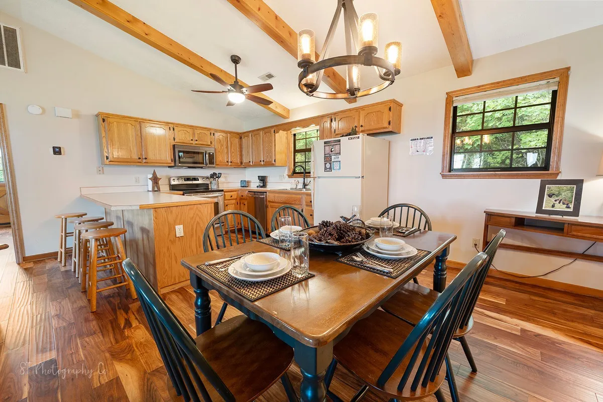 A close-up of the dining table set for a meal, with rustic wooden beams overhead and warm lighting. The background reveals a well-equipped kitchen with cabinetry and modern appliances, inviting guests to cook and dine together.