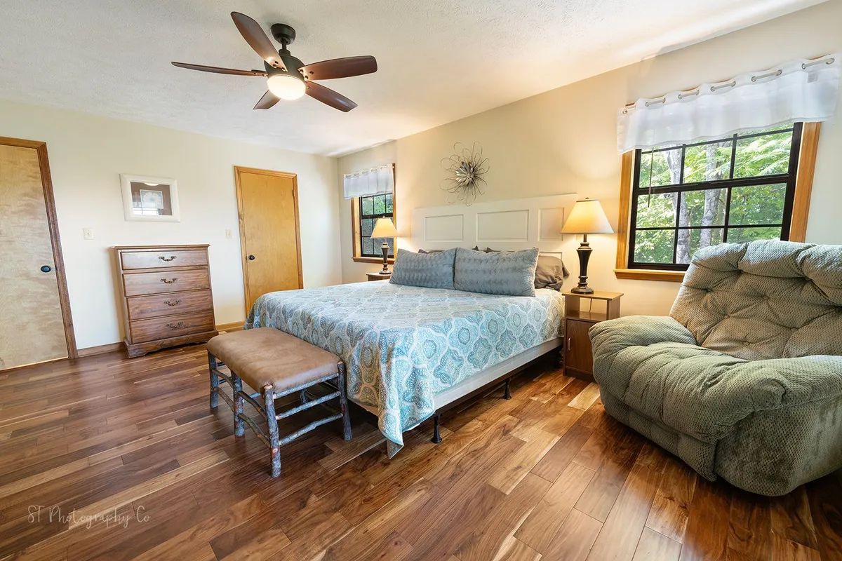 A comfortable king bedroom outfitted with decorative bedding, a recliner chair for reading or relaxing, and a wooden dresser for ample storage. Sunlight streams into the room, creating a cozy and tranquil atmosphere ideal for unwinding.