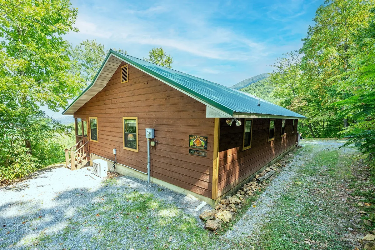 This image captures the exterior of the cabin with welcoming wooden siding, a green metal roof, and a pathway leading to the entrance, all set amidst lush, mature trees. The peaceful setting and the inviting porch highlight the cabin's connection to nature and its potential as a launching point for adventure.