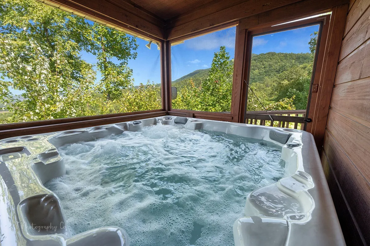 This image features a tranquil interior scene with a hot tub positioned beneath rustic wooden beams, offering scenic views of the mountains through a large window. The inviting space suggests relaxation, socializing, and the enjoyment of exclusive amenities within the cabin.