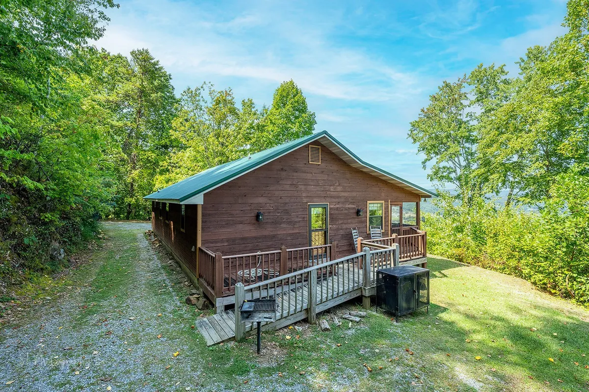 This image shows the inviting exterior of a rustic wooden cabin set amid lush trees. A spacious deck wraps around the structure, offering plenty of outdoor seating and scenic views—ideal for sipping wine, relaxing at sunset, or savoring the peaceful mountain atmosphere.