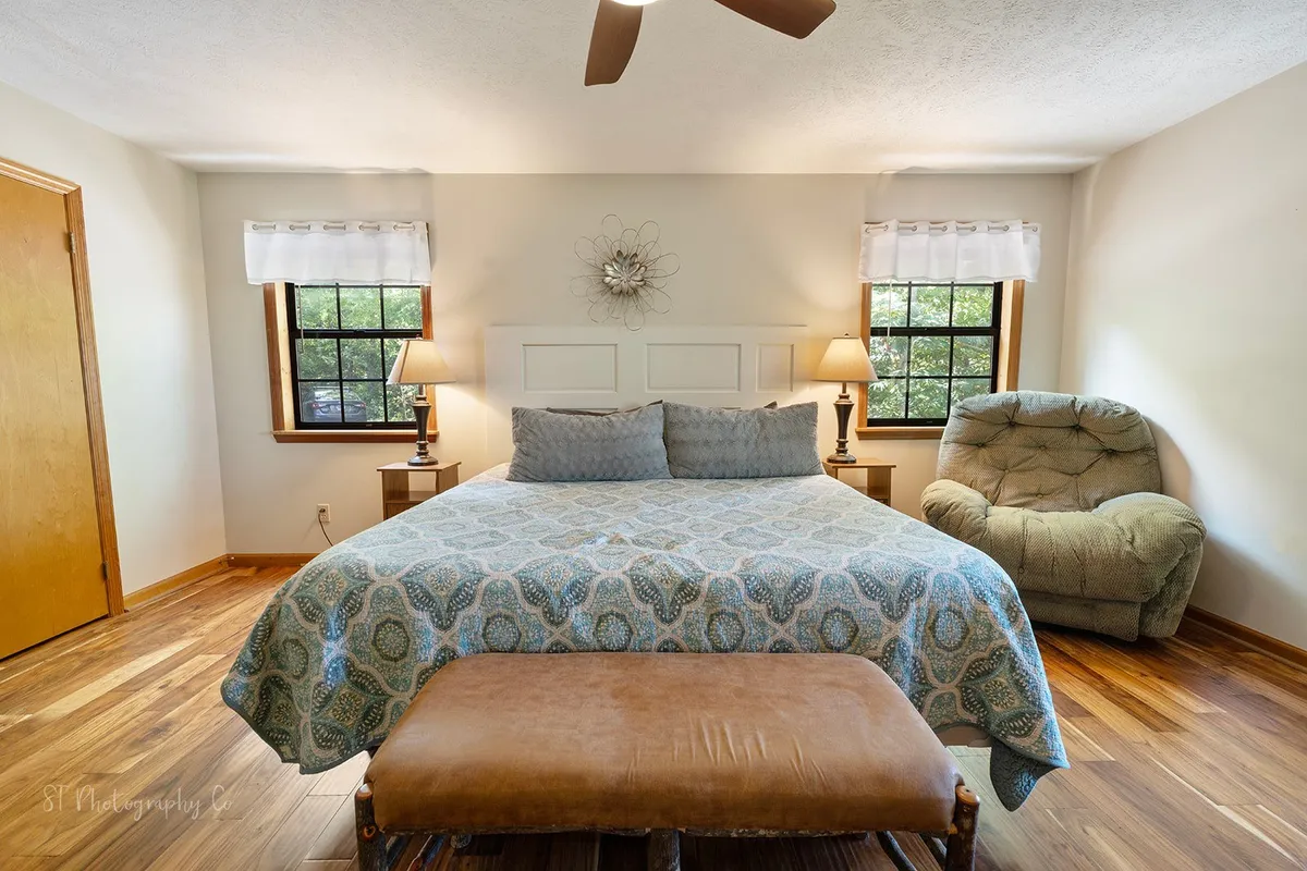 A serene bedroom with a large bed dressed in patterned bedding, a wooden bench, a comfortable armchair, and a decorative wall piece. The layout is inviting, ideal for peaceful mornings with coffee or cozy evenings with a book.