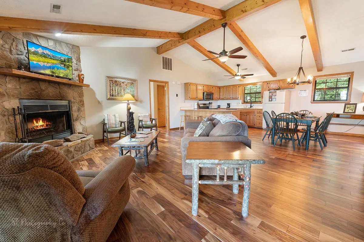 A spacious, warmly lit living room featuring a plush sofa and armchairs circling a robust wood-burning fireplace. Beyond the seating area, a dining table is set for a meal, and a flat-screen TV displays a scenic image. Large windows let in natural light and frame the view of the mountains outside.